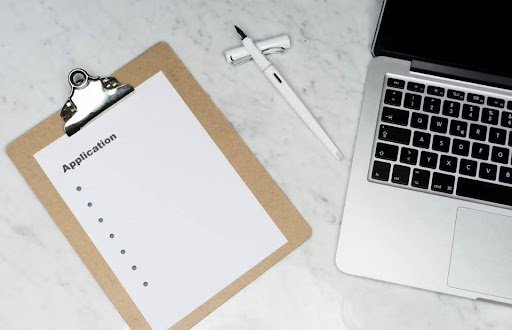 Application on clip board set near a laptop and pen. Photo by Unsplashed.com.